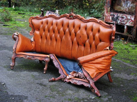 An outdoor scene featuring a vintage-style sofa with a wooden carved frame and tufted upholstery in a warm, brown leather-like material. The sofa is damaged, with noticeable tearing and missing sections on the right armrest and lower side, exposing the internal padding and supporting structure. It is positioned on a paved surface, likely part of a driveway or patio area, with a background of greenery including grass and trees, and a section of weathered building or shed with graffiti visible in the upper right corner. The overall environment appears unkempt, consistent with a rubbish or waste removal context. This image highlights the need for professional waste removal services such as those offered by Commercial Waste Southwark, specialising in the collection and disposal of large household furniture and vintage items in the Southwark area, near postcode SE1 or SE5.