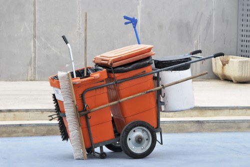 A professional waste management trolley fitted with two large black rubber wheels and a sturdy metal frame, positioned outdoors on a paved surface adjacent to a concrete wall. The trolley is orange and features a large rectangular plastic container T-shaped, with a lid secured on top and several long-handled tools, including a broom with a white bristle head, a hand rake, and a dustpan, leaning against or attached to the trolley. The background includes a concrete step and a small section of a modern building with a light grey and metallic finish, indicating an urban or commercial environment, possibly in Southwark. The scene reflects a setting ready for rubbish or waste collection, aligned with commercial waste removal services like those offered by Commercial Waste Southwark.