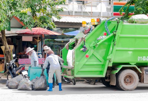 A green refuse collection truck with a metallic finish is parked on a city street, positioned parallel to the pavement. The truck has a large, enclosed cargo area with a hydraulic lifting mechanism at the rear, which is in use collecting waste. Two sanitation workers wearing light grey uniforms, protective blue gloves, and white helmets are actively engaged in emptying black rubbish bags onto the truck's intake point; one worker is bending forward while the other guides the bags. The scene includes several overfilled black rubbish bags on the ground in front of the workers. In the background, there are small shops or market stalls, some with colorful umbrellas providing shade, and trees with green foliage lining the street. The overall environment indicates a typical urban setting, possibly in South London, where Commercial Waste Southwark operates, with natural lighting suggesting daytime.