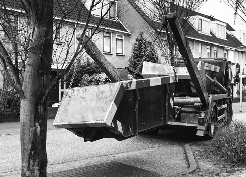 A black flatbed tow truck parked on a residential street in Southwark, with a large, rectangular metal container filled with various types of waste materials secured on its platform. The container appears to be made of weathered, light-colored metal with visible rust and dirt, reflecting typical roadside rubbish collection equipment. The truck's hydraulic lifting arm is raised, supporting the container, and is positioned near the right side of the vehicle. The background features rows of terraced houses with pitched roofs, some with dormer windows, and leafless trees indicating a colder season. The street is paved with asphalt, and a concrete sidewalk runs alongside it, with some shrubs present along the front gardens of the houses. The overall scene portrays active rubbish removal or waste collection, consistent with services provided by Commercial Waste Southwark to support local businesses and residents in the area.