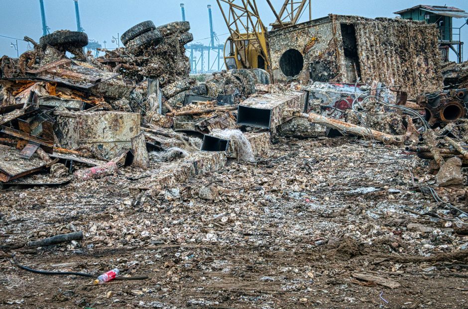 A large, cluttered pile of rusted, weathered scrap metal and debris situated outdoors, with various metal sheets, pipes, and chassis parts scattered across a rough, uneven ground of dirt and small rub