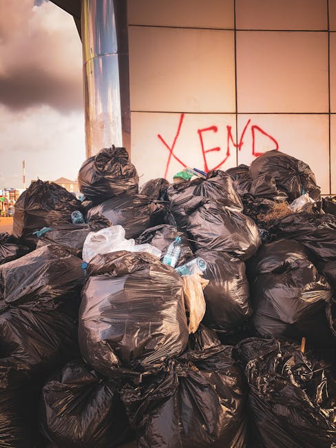 A narrow urban alleyway filled with a large, rolling waste bin in the foreground, positioned on a dirty, uneven pavement. The bin appears to be made of weathered, grey plastic or metal, and is slightl