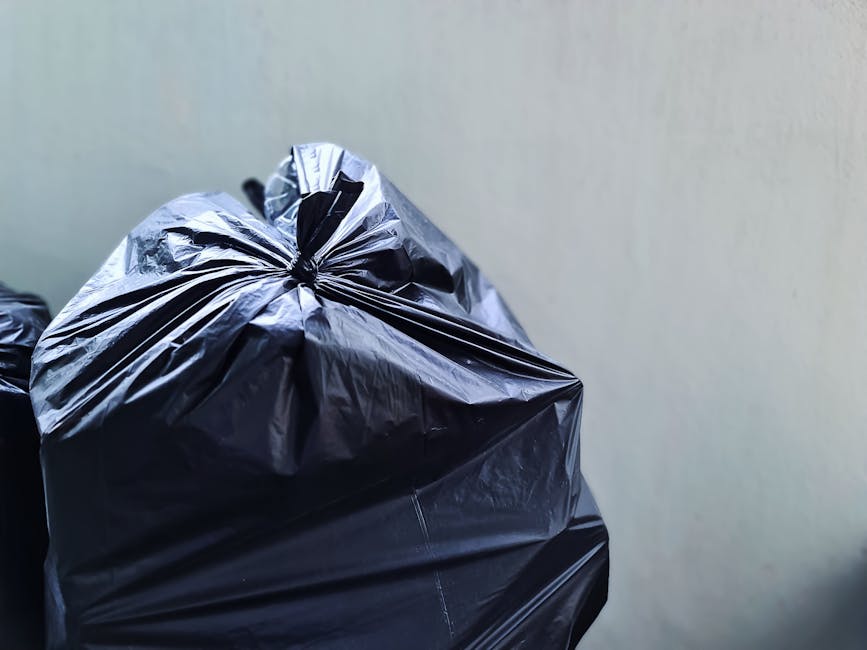 Close-up of several large black plastic rubbish bags filled with waste, stacked against a plain light-colored wall. The bags are made of shiny, crinkled plastic material, with some tied at the top to 